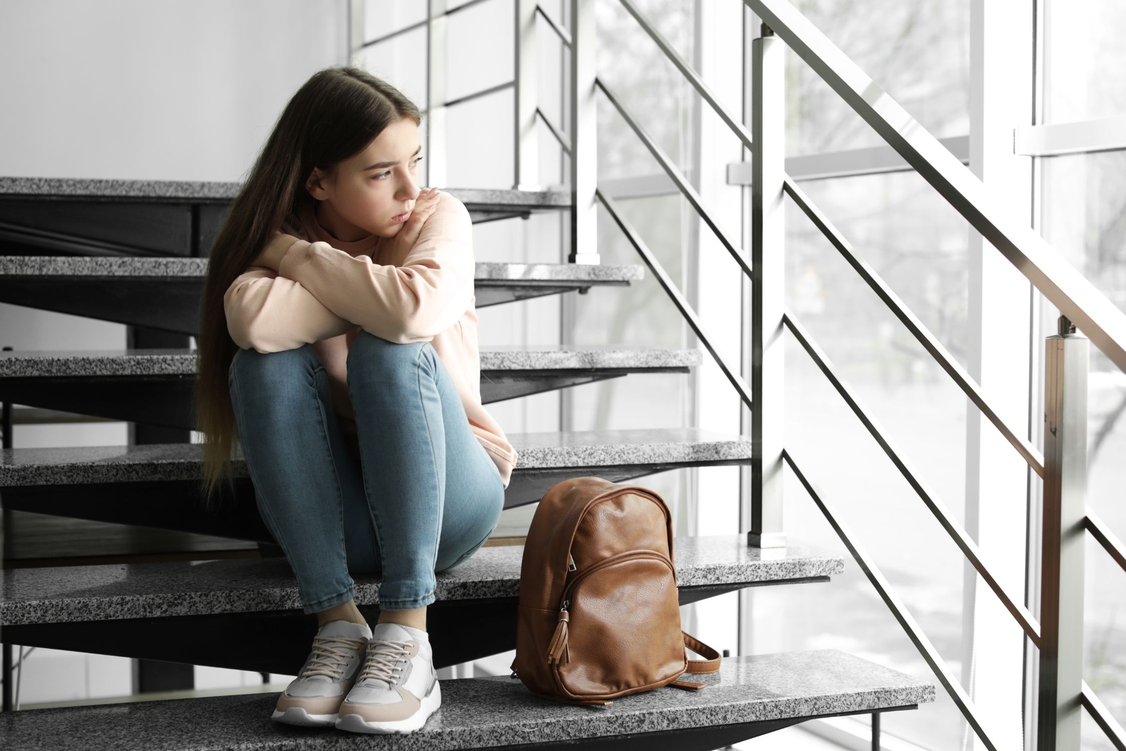 Jugendliche sitzt mit auf den Knien verschräbkten Armen auf einer Treppe und schaut traurig aus dem Fenster