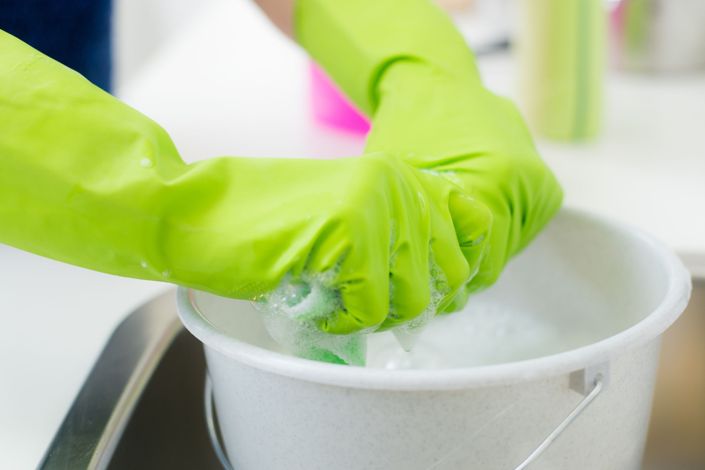 Hände in grünen Gummihandschuhen wringen einen nassen Lappen über einem Eimer aus