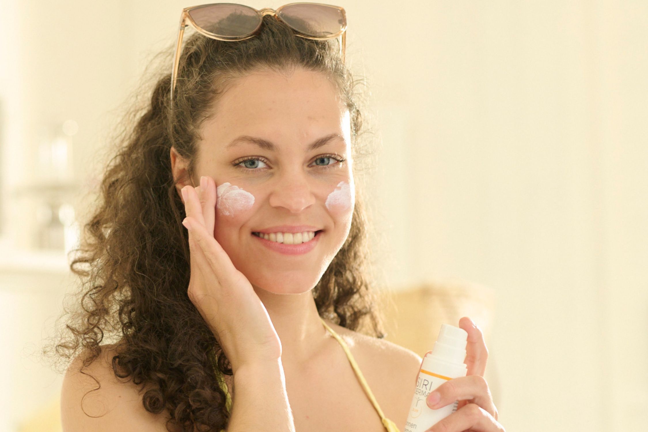 Eine Frau mit dunklen Locken und Sonnenbrille im Haar cremt sich die Wangen mit Sonnencreme ein